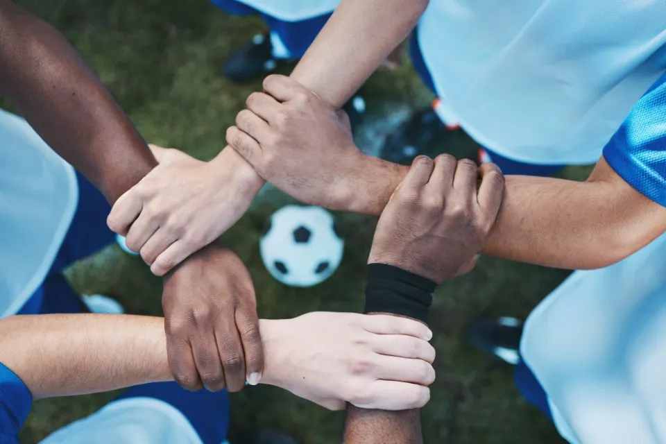Beneficio de integrar actividades deportivas como el Mundialito: mejorar la cohesión en los equipos de trabajo. Mundialito: fotografía de 5 personas uniendo su brazos y manos formando un cuadrado y al centro de este un balón de fútbol.