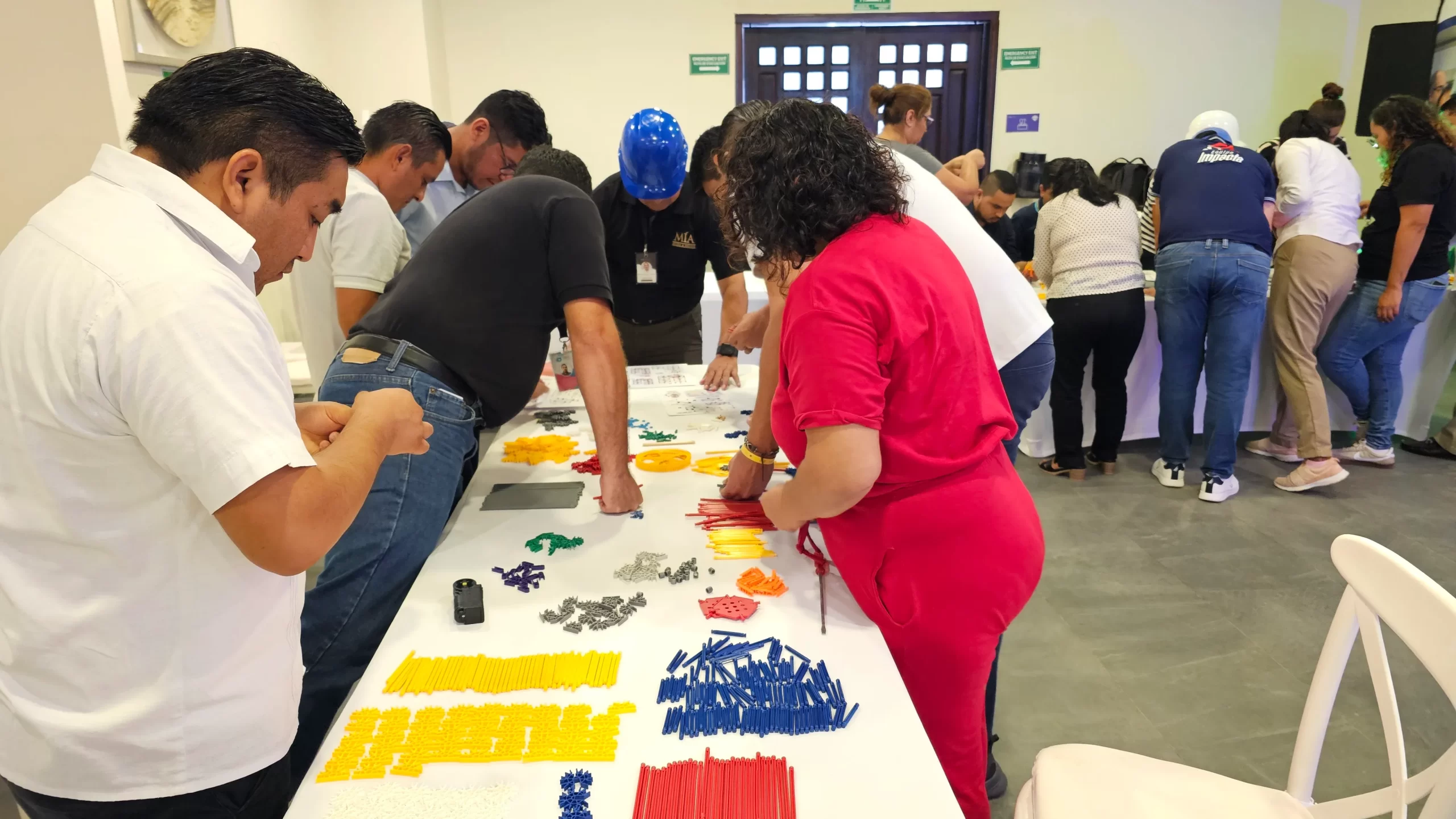 Ejemplo de las actividades de team building híbrido que se realizan para fortalecer la relación de un equipo de trabajo. Team building híbrido: Fotografía de personas alrededor de una mesa con mantel color blanco utilizando figuras de colores.
